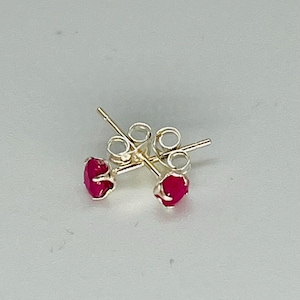 May include: A pair of small stud earrings featuring bright red gemstones. The earrings have silver-coloured posts and backs. The gemstones are round and polished. The earrings are displayed on a light grey surface.