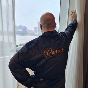 May include: A black robe with the name "Roman" embroidered on the back in gold thread. The robe is worn by a person standing in front of a window with a view of a city skyline.