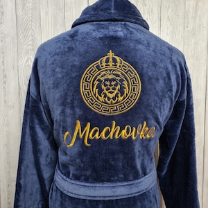 Puede incluir: Una bata azul marino con un diseño de cabeza de león bordado en oro dentro de un círculo con una corona sobre la cabeza del león. El texto "Machovka" está bordado debajo del diseño de la cabeza del león.