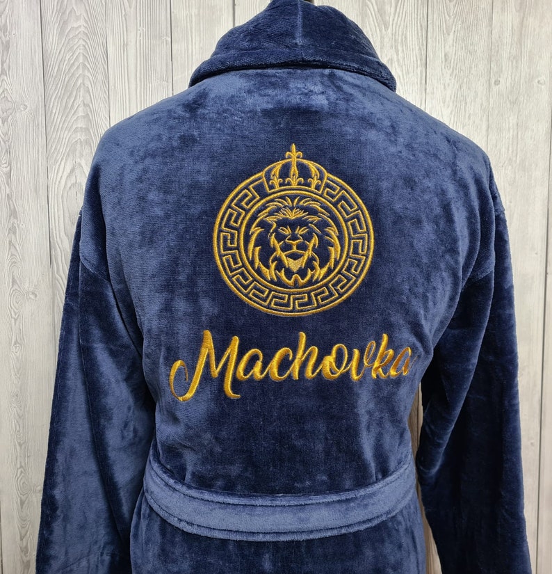 K&ouml;nnte beinhalten: Ein marineblauer Bademantel mit einem goldenen gestickten L&ouml;wenkopf-Design in einem Kreis mit einer Krone &uuml;ber dem L&ouml;wenkopf. Der Text "Machovka" ist unterhalb des L&ouml;wenkopf-Designs gestickt.