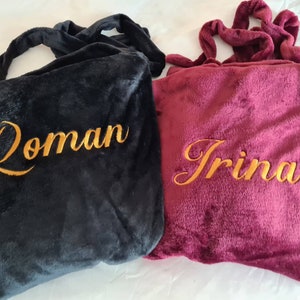 May include: Two personalized fleece blankets, one black with the name "Roman" embroidered in gold, and one burgundy with the name "Irina" embroidered in gold.