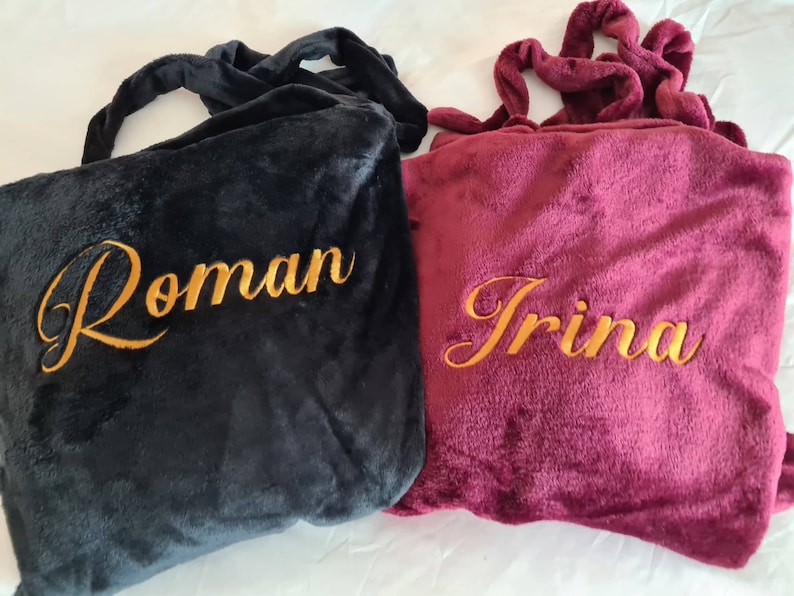May include: Two personalized fleece blankets, one black with the name "Roman" embroidered in gold, and one burgundy with the name "Irina" embroidered in gold.