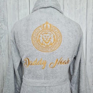 K&ouml;nnte beinhalten: Graue Kapuzenrobe mit einem goldenen gestickten L&ouml;wen- und Kronendesign. Der Text "Daddy Noah" ist unter dem L&ouml;wen gestickt.