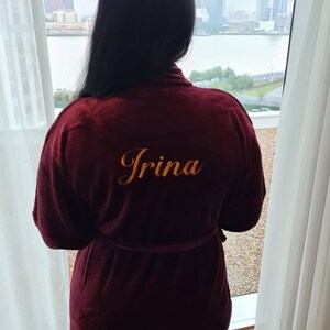 May include: A maroon robe with a gold embroidered name "Irina" on the back. The robe is tied at the waist and the person wearing it is looking out a window.