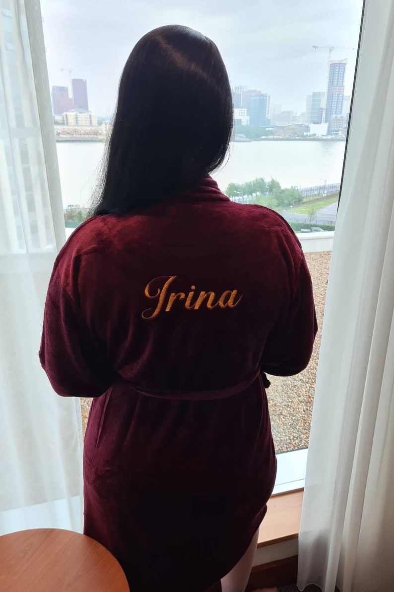 May include: A maroon robe with a gold embroidered name "Irina" on the back. The robe is tied at the waist and the person wearing it is looking out a window.