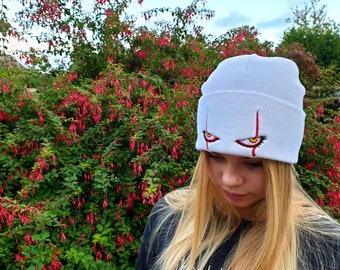 May include: White beanie with red and yellow embroidered eyes. The eyes are wide open and have a menacing look.