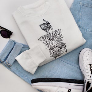 May include: White sweatshirt featuring a black and white illustration of a cat gazing into a pool of water, reflecting a tiger. The sweatshirt is folded and placed on a white surface with blue jeans and white trainers.