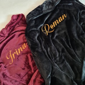 May include: Two plush robes with hoods, one burgundy and one black, with gold embroidery. The burgundy robe has the name "Irina" embroidered on it, and the black robe has the name "Roman" embroidered on it.