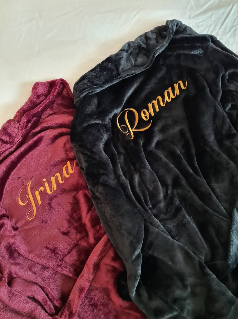 May include: Two plush robes with hoods, one burgundy and one black, with gold embroidery. The burgundy robe has the name "Irina" embroidered on it, and the black robe has the name "Roman" embroidered on it.