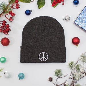 May include: A dark grey beanie with a white peace sign embroidered on the front. The beanie is surrounded by Christmas decorations and greenery.
