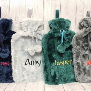 May include: Four personalised faux fur hot water bottles in navy blue, grey, green, and grey with pom poms. The names Theo, Amy, Jasper, and Mansi are embroidered on the bottles.