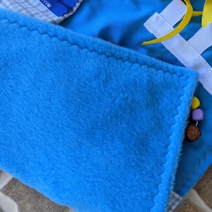 May include: A blue fleece fabric square with a zig-zag stitch around the edge. The fabric is part of a larger multi-textured activity mat.