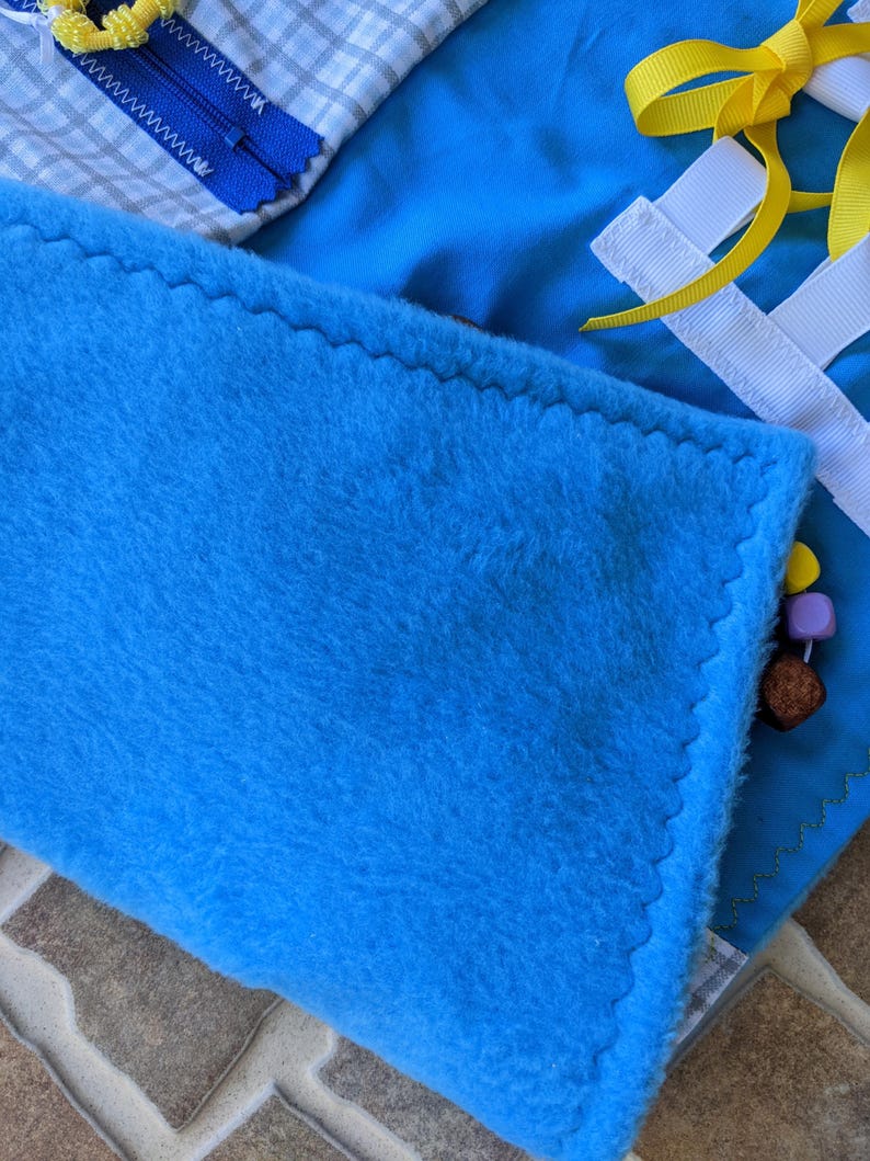 May include: A blue fleece fabric square with a zig-zag stitch around the edge. The fabric is part of a larger multi-textured activity mat.