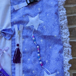 May include: A purple and white sensory blanket with a zipper, a star, a beaded string, and a tic-tac-toe board. The blanket has a white lace trim and a white strap with a buckle.