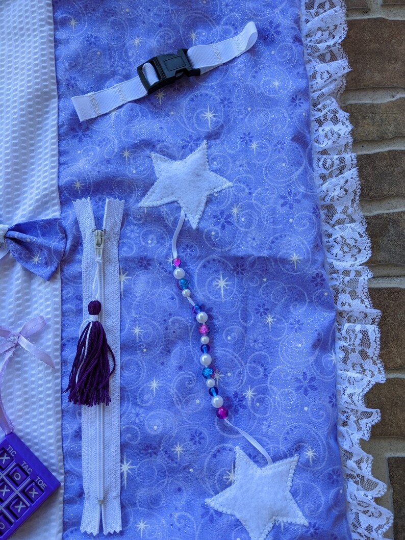 May include: A purple and white sensory blanket with a zipper, a star, a beaded string, and a tic-tac-toe board. The blanket has a white lace trim and a white strap with a buckle.