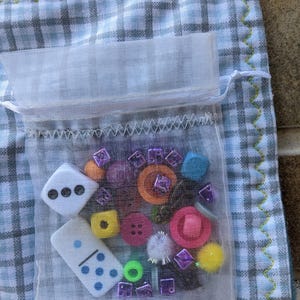 May include: A white mesh bag filled with colorful buttons, dice, and other small trinkets. The bag is sitting on a blue and white checkered fabric.