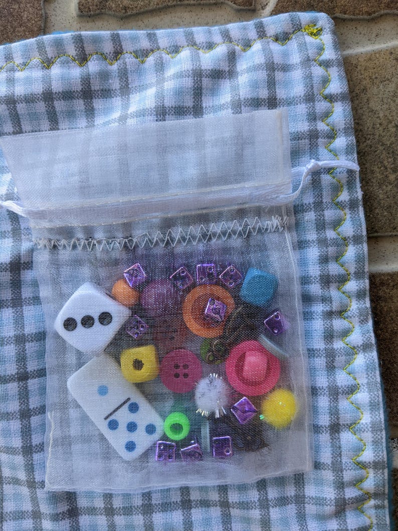 May include: A white mesh bag filled with colorful buttons, dice, and other small trinkets. The bag is sitting on a blue and white checkered fabric.