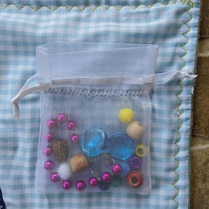 May include: A white mesh bag filled with colorful beads, buttons, and other small objects. The bag is sitting on a blue and white checkered fabric.