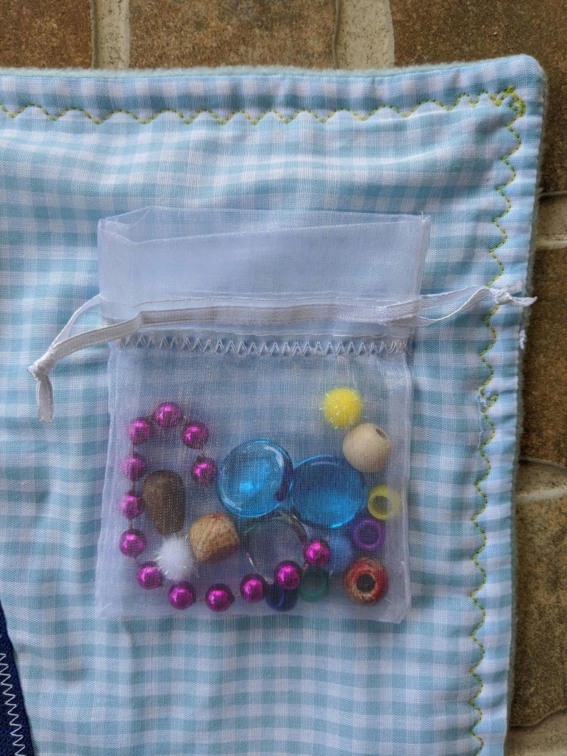 May include: A white mesh bag filled with colorful beads, buttons, and other small objects. The bag is sitting on a blue and white checkered fabric.