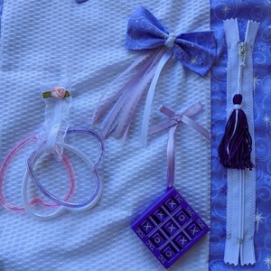 May include: A white fabric square with a purple tic-tac-toe game, a purple bow with white and pink ribbons, a purple zipper with a tassel, and two pink and purple heart-shaped rings.