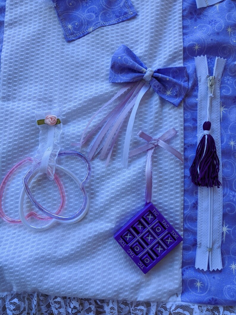 May include: A white fabric square with a purple tic-tac-toe game, a purple bow with white and pink ribbons, a purple zipper with a tassel, and two pink and purple heart-shaped rings.