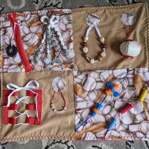 May include: A beige and brown fabric square with different textures and activities for babies and toddlers. The square is divided into four sections, each with a different activity. The top left section has a zipper, a white and gray tassel, and a red and white baseball pattern. The top right section has a white and brown beaded necklace, a white string with a knot, and a white baseball with a red ribbon. The bottom left section has a red and white lacing activity, a white string with a knot, and a white and gray beaded necklace. The bottom right section has a red and white baseball pattern, a blue and orange string with beads, and a red and white string with beads.