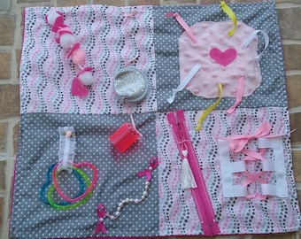 May include: A pink and gray baby activity mat with various textures and objects for sensory play. The mat features a zipper, a heart-shaped patch, a plastic container, a string of pearls, and a variety of ribbons.