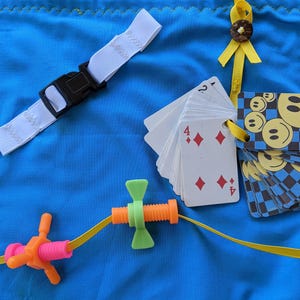 May include: A blue fabric square with a white strap, a yellow ribbon with a button, a deck of playing cards with the number 4 of diamonds showing, and a toy with a screw and a butterfly-shaped piece.