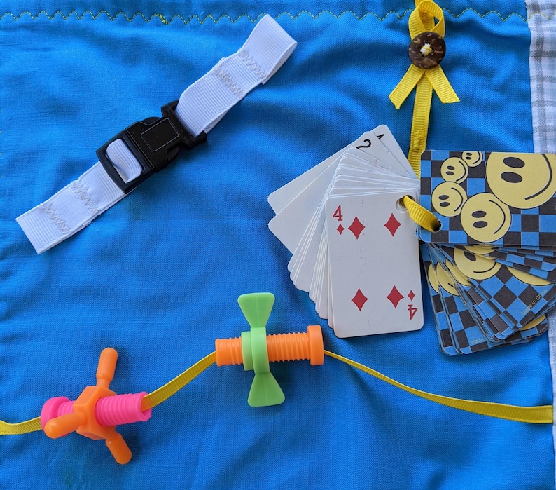 May include: A blue fabric square with a white strap, a yellow ribbon with a button, a deck of playing cards with the number 4 of diamonds showing, and a toy with a screw and a butterfly-shaped piece.