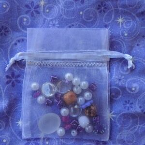 May include: A white organza bag filled with a variety of small, colorful beads, charms, and other decorative items. The bag is on a blue fabric background with a white star pattern.