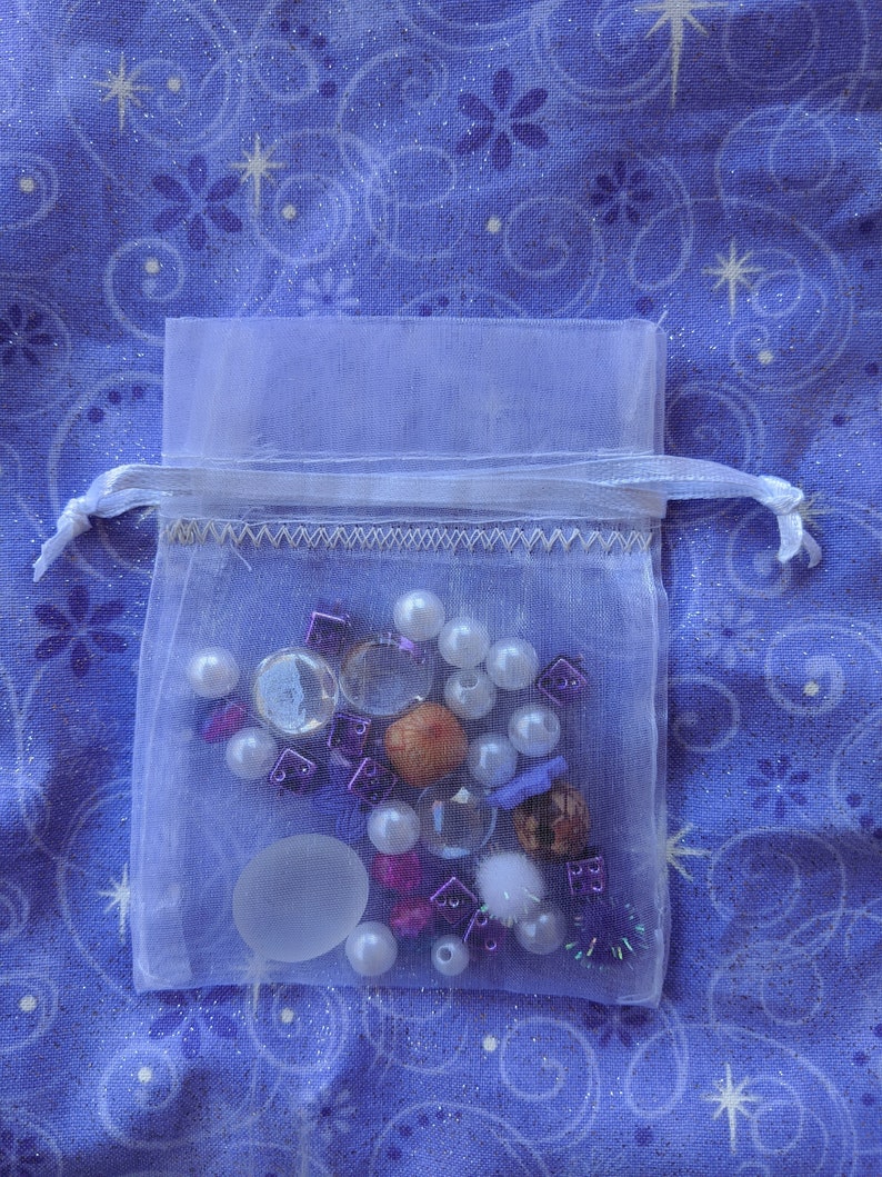 May include: A white organza bag filled with a variety of small, colorful beads, charms, and other decorative items. The bag is on a blue fabric background with a white star pattern.