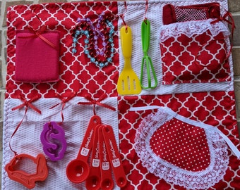 May include: A red and white fabric play mat with various textures and shapes. The mat includes a red fabric square with a white lace trim, a red fabric square with white polka dots, a white fabric square with a red fabric heart, a red fabric square with a white lace trim, and a white fabric square with a red fabric heart. The mat also includes a set of measuring spoons, a set of cookie cutters, a set of kitchen utensils, and a set of beads.