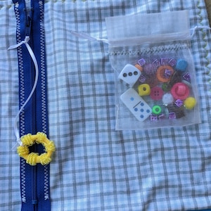 May include: A blue and white checkered fabric pouch with a zipper closure. The pouch has a small mesh bag attached to it, filled with colorful buttons, beads, and dice.
