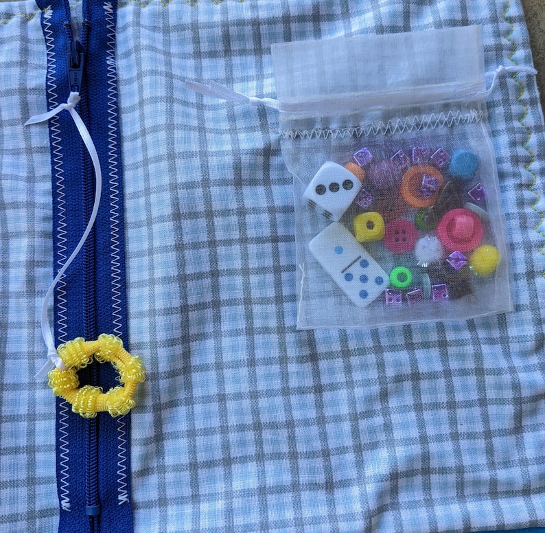May include: A blue and white checkered fabric pouch with a zipper closure. The pouch has a small mesh bag attached to it, filled with colorful buttons, beads, and dice.