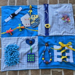 May include: A blue and white sensory play mat with various textures and activities for children. It features a zipper, a tic-tac-toe board, a heart-shaped button, a fluffy blue piece, a string of beads, a ribbon, a plastic toy, a set of playing cards, and a small clear bag with dice and beads.