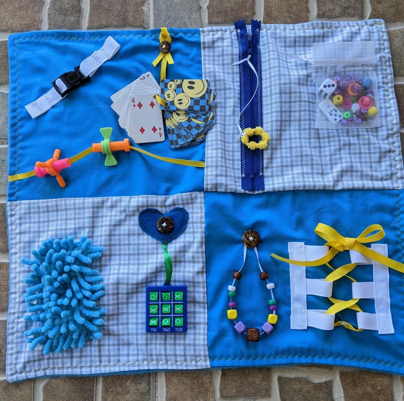 May include: A blue and white sensory play mat with various textures and activities for children. It features a zipper, a tic-tac-toe board, a heart-shaped button, a fluffy blue piece, a string of beads, a ribbon, a plastic toy, a set of playing cards, and a small clear bag with dice and beads.