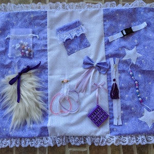 May include: A purple and white sensory blanket with various textures and activities for babies and toddlers. It features a zipper, a fuzzy piece, a small pouch with beads, a bow, a star, a small pouch with a zipper, a hair clip, a small pouch with a zipper, and a small plastic toy.