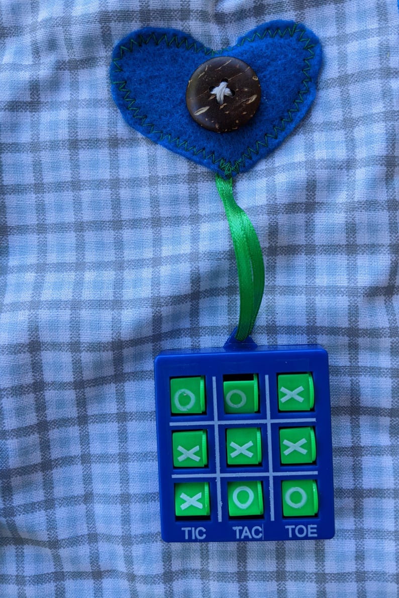 May include: A blue and green tic-tac-toe game with a blue heart-shaped fabric ornament with a brown button on top. The ornament is hanging from a green ribbon.