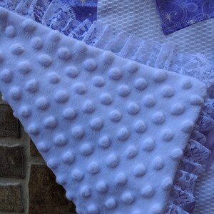 May include: A soft, lavender-colored baby blanket with a textured, dotted surface. The blanket is trimmed with white lace and has a section of purple fabric with a star pattern.