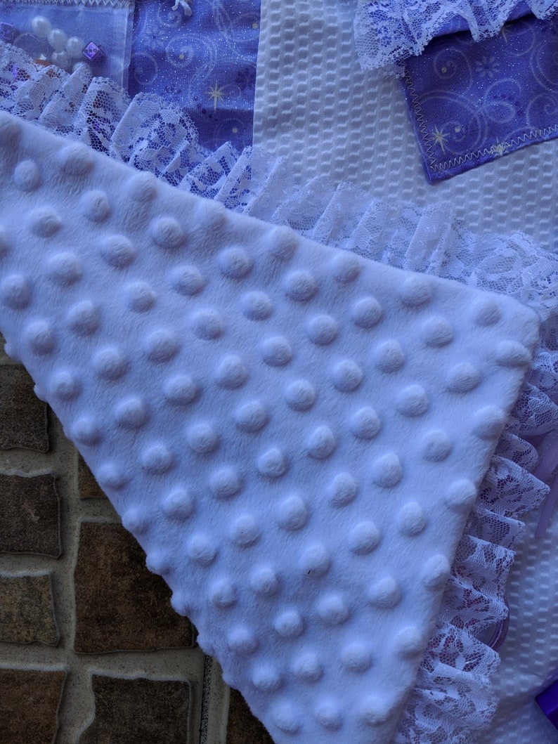 May include: A soft, lavender-colored baby blanket with a textured, dotted surface. The blanket is trimmed with white lace and has a section of purple fabric with a star pattern.