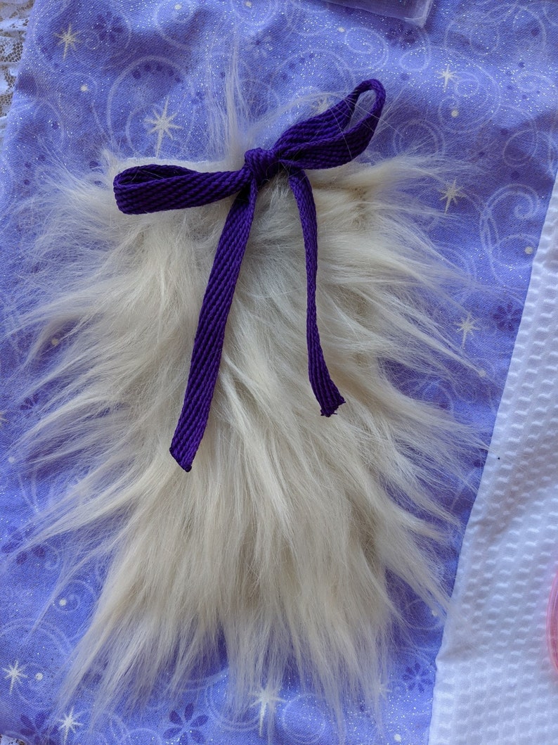 May include: A white fluffy tail with a purple ribbon tied around it. The tail is on a purple fabric with a white star pattern.