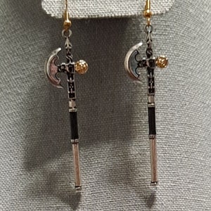 May include: A pair of silver and gold axe earrings. The axe heads are silver with black detailing and the handles are gold with black detailing.