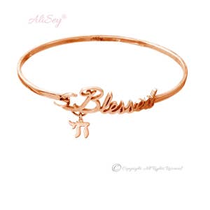 May include: A rose gold bangle bracelet with the word "Blessed" inscribed on it. A charm with the Hebrew letter "Chai" hangs from the bracelet.