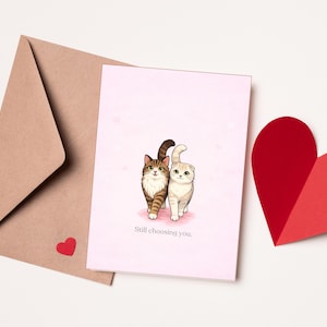 Pode incluir: Um cartão de felicitações rosa com uma ilustração de dois gatos e o texto "Still choosing you". Um envelope castanho e dois autocolantes vermelhos em forma de coração estão à esquerda. Um grande coração vermelho está à direita.