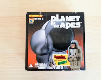 Könnte beinhalten: Eine schwarze und orangefarbene Schachtel mit einem weißen Bild eines Affen, der einen Raumanzug trägt. Auf der Schachtel steht "Planet der Affen" und "Cornelius als Astronaut mit Ikarus im Inneren".
