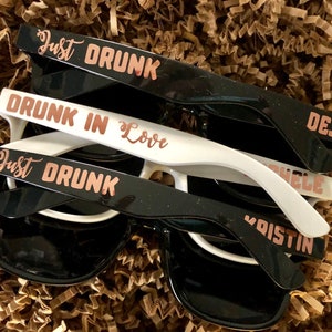 May include: A set of four black and white plastic sunglasses with rose gold lettering. The sunglasses have the following text: "Just Drunk", "Drunk in Love", "Deja", and "Just Drunk".