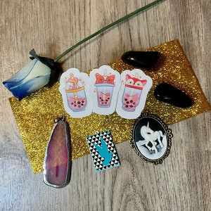 May include: An assortment of items on a wooden surface, including stickers of bubble tea with animal faces, a pendant with a pink stone, a black and white unicorn cameo, and black stones. A blue rose and gold glitter paper are also present.