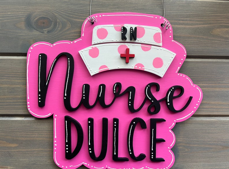 Peut inclure: Une pancarte en bois rose avec des pois blancs et le texte "Nurse Dulce" en lettres noires. Un bonnet d'infirmi&egrave;re blanc avec des pois roses et les lettres "RN" en noir est plac&eacute; sur le dessus de la pancarte.