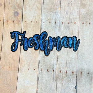 May include: Blue and black glitter letters spelling out the word "Freshman" on a wood background.