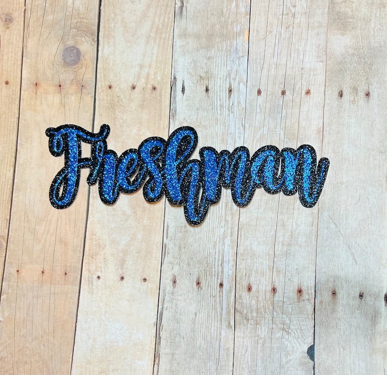 May include: Blue and black glitter letters spelling out the word "Freshman" on a wood background.